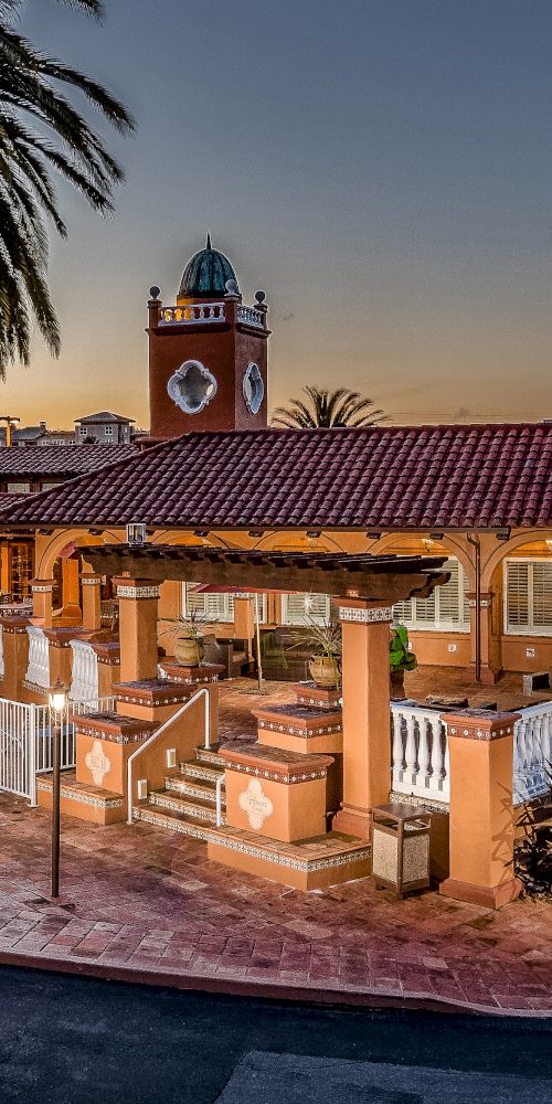 The image shows a Spanish-style building with a clock tower, surrounded by palm trees and featuring a pool area. The scene is illuminated at dusk.