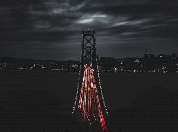 A nighttime photo of a bridge with car light trails, against a backdrop of cloudy skies and a distant cityscape with illuminated buildings.