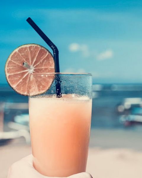 A hand holding a glass of juice with a slice of lime and a black straw, set against a background of a beach and the sea.