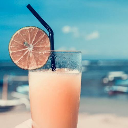 A hand holding a glass of juice with a slice of lime and a black straw, set against a background of a beach and the sea.
